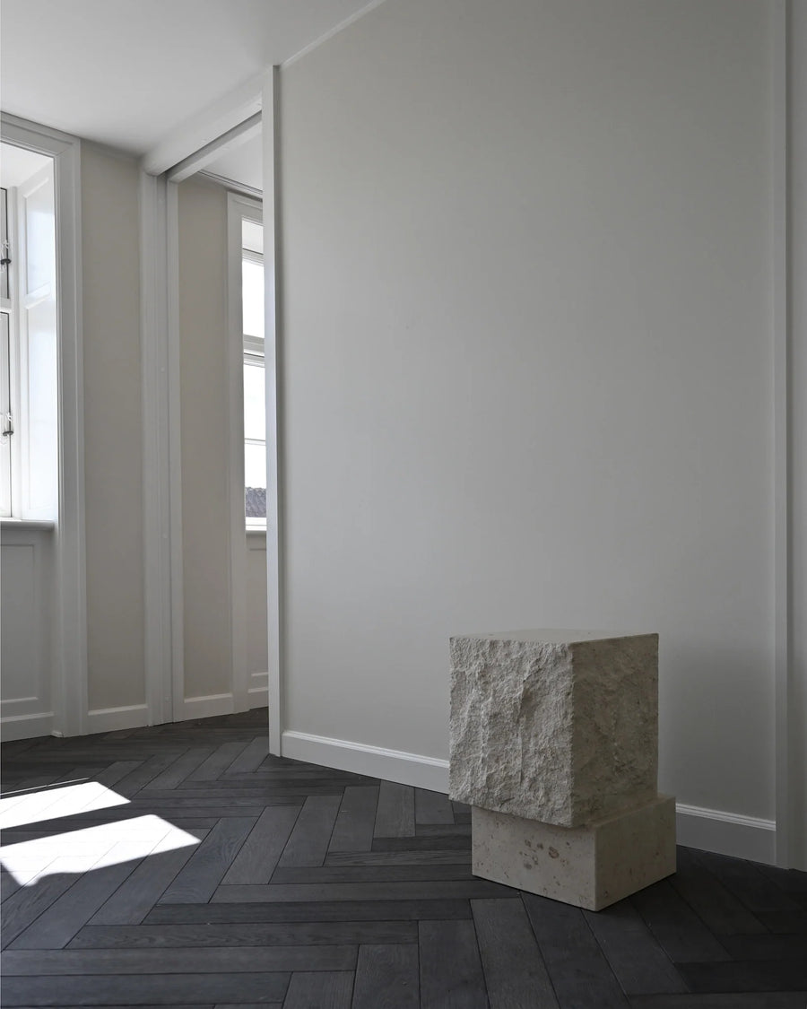 Tall limestone plinth in a minimalistic room with white walls and wooden floor.