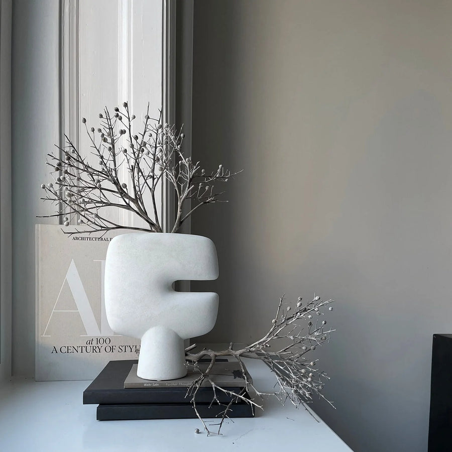 White abstract vase with branches on a surface near a window styled with coffee table books. 