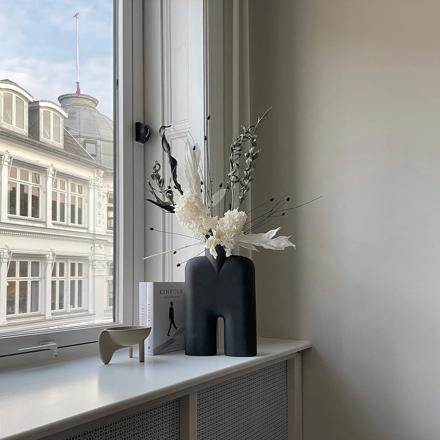 Decorative vase with flowers on a windowsill with a view of a building outside.