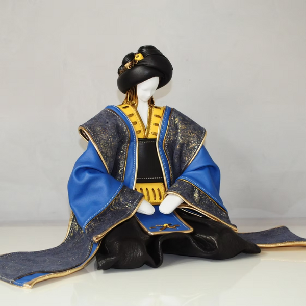 A traditional Japanese leather figurine wearing a blue and black kimono with gold trim, seated against a plain light gray background.