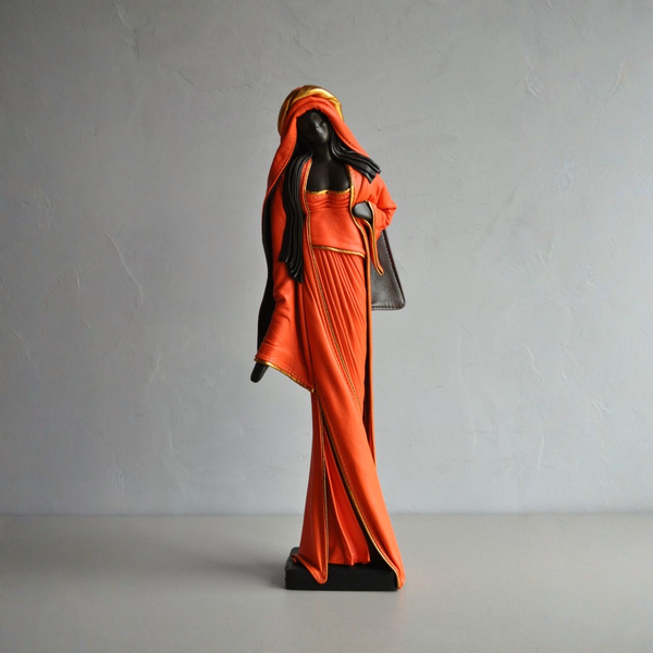 A decorative figurine of a woman in a flowing orange robe and headscarf with black hair and black base details, standing against a light gray textured wall.