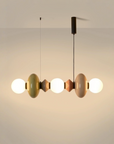 A modern multi-light linear pendant chandelier with glossy, rounded bulb-shaped shades in muted olive green, cream, and terracotta, suspended by black cords against a warm beige wall.