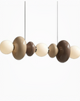 A modern multi-light linear pendant chandelier with glossy, organic-shaped bulbs in beige, brown, and cream, suspended by thin black cords against a plain white background.