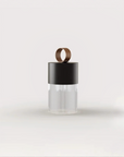A modern cylindrical portable and rechargeable with a matte black upper section, a ribbed clear lower section, and a small metallic bronze ring handle on top, centered against a light gray background.