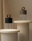 Two modern cylindrical portable and rechargeable lights with ribbed stone-look pedestals, one in black and one in taupe, each with a clear glass lower section and a brown leather ring handle, set against a warm beige background.