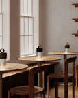 A modern room with round wooden tables and chairs, featuring three portable LED table lamps with black and metallic caps placed on the tabletops. The background includes a warm beige wall, frosted window panes, wooden shelves, and a wooden bench.