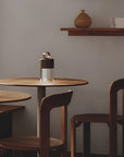 A modern portable and rechargeable table lamp with a clear cylindrical body and a dark brown top sits on a round wooden table in a minimalist dining setting with matching wooden chairs and a shelf in the background.