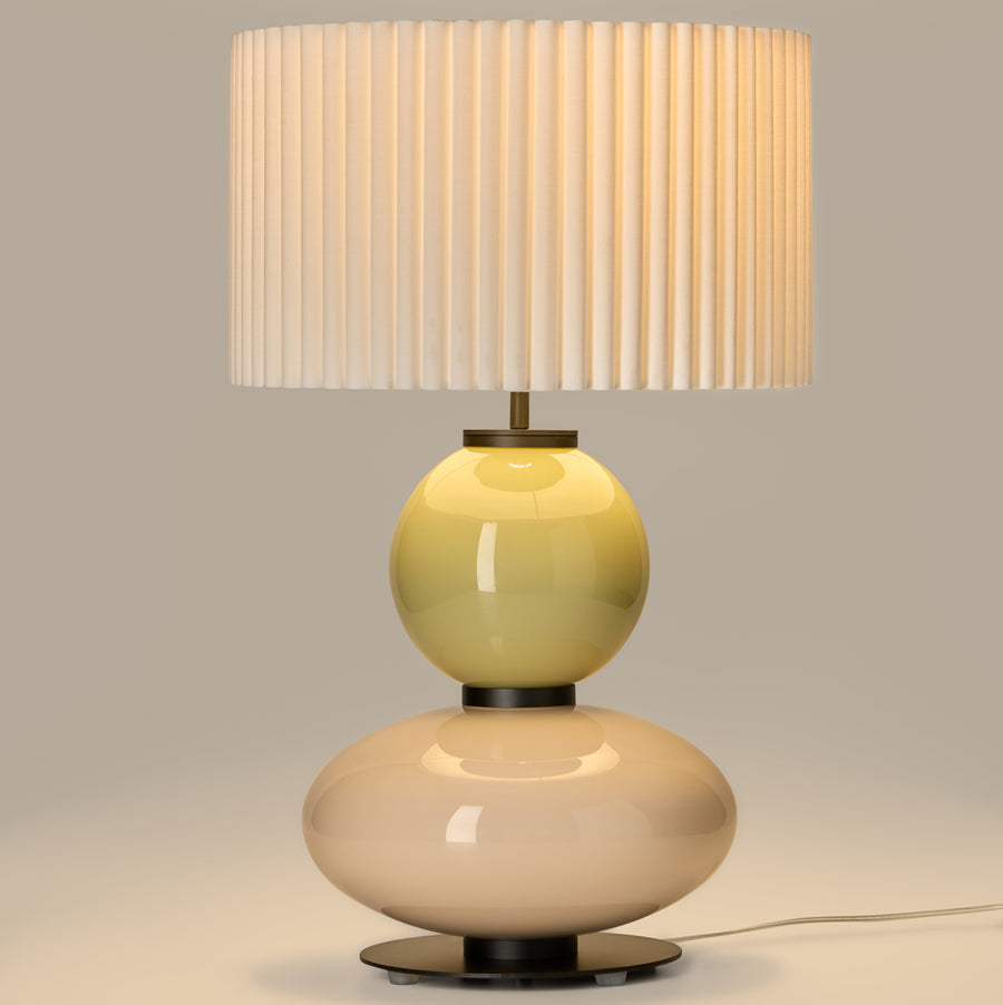 Lit up, modern table lamp with a yellow and white glass base and a pleated shade from Aromas. Available at Spacio retails stores in India. 
