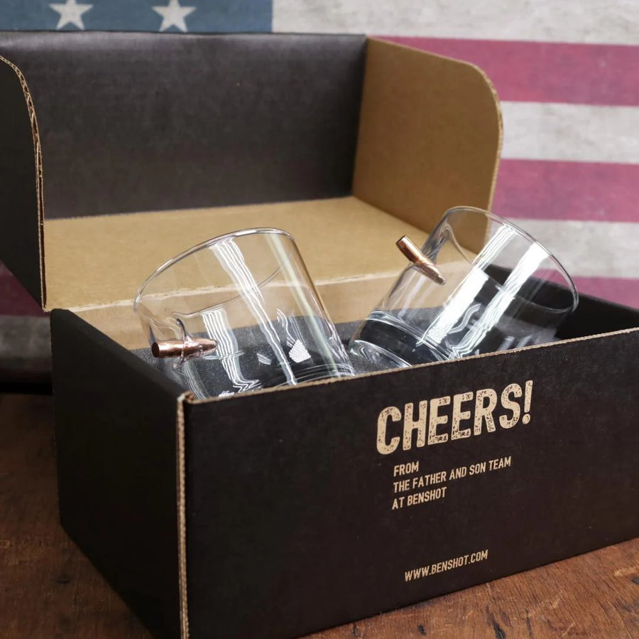 Clear glass bullet-shaped shot glasses in a black box with 'Cheers!' text against an American flag background.