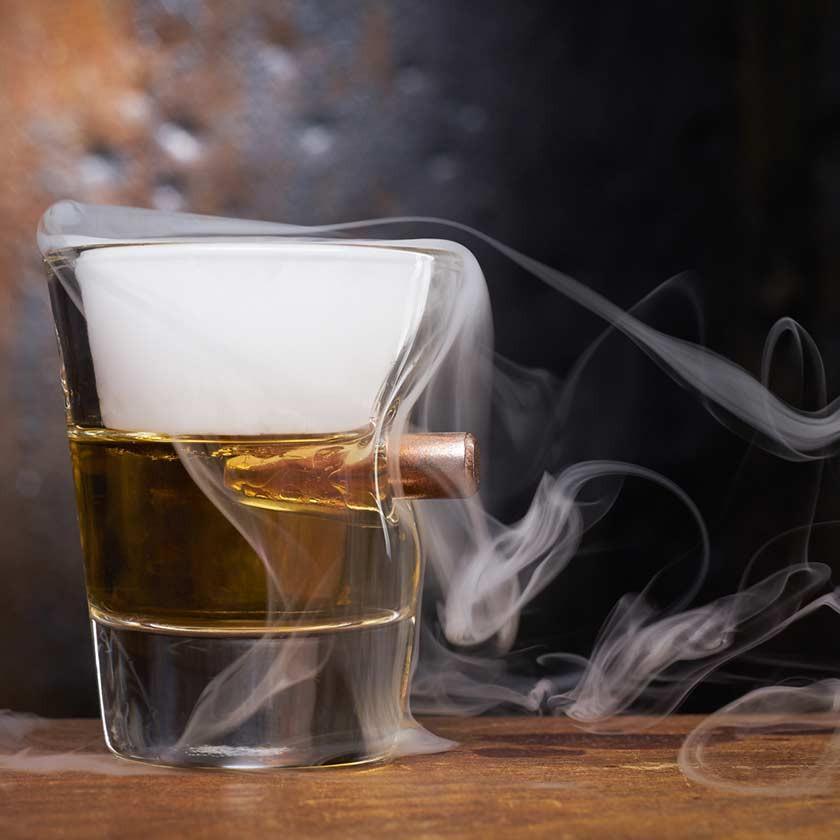 Glass with a bullet and liquid, surrounded by smoke on a wooden surface.