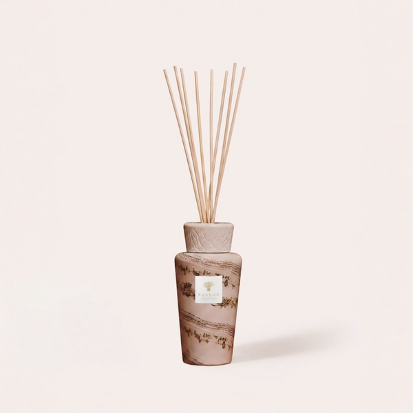 A reed diffuser bottle with a textured beige cap and natural wood reeds, featuring a brown and gold botanical patterned glass body with a white label, set against a solid cream background.