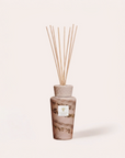 A reed diffuser bottle with a textured beige cap and natural wood reeds, featuring a brown and gold botanical patterned glass body with a white label, set against a solid cream background.