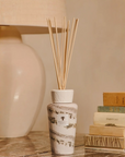 A reed diffuser bottle with natural wood reeds, decorated with a botanical pattern, sits on a marble surface next to a stack of vintage books and a large ceramic lamp in the background.