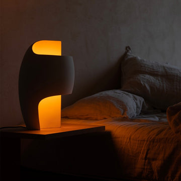 Modern table lamp with a warm glow on a bedside table in a dimly lit room.
