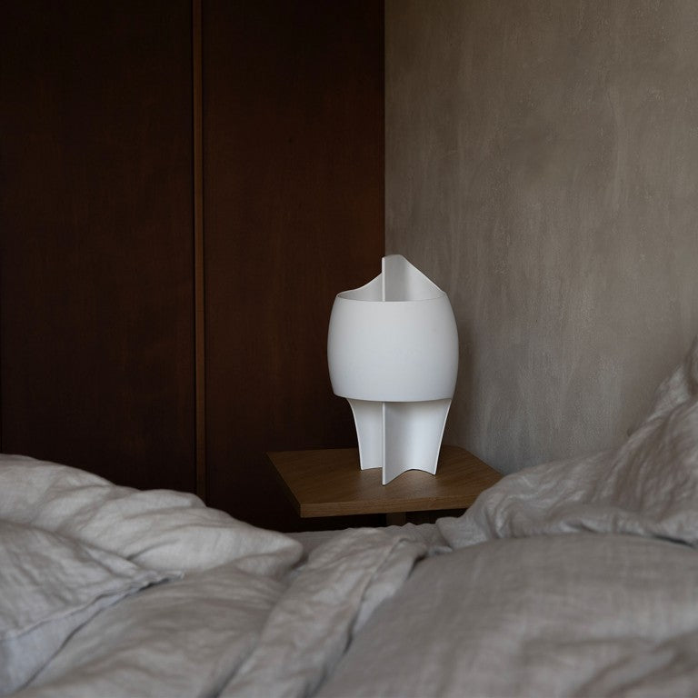 White sculptural table lamp on a wooden shelf next to a bed with gray bedding.