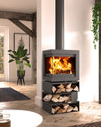 A modern black freestanding wood-burning fireplace with a visible fire inside, set in a styled living room with wood floors, rustic beams, and a plant.