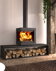 A modern black wood-burning fireplace with a visible fire, mounted on a matching base shelf holding stacked firewood, in a styled interior room with wood flooring, a chair, and a tree trunk.