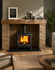 A black wood-burning fireplace with a glass front and active fire, set within a brick fireplace surround with a rustic wooden mantel in a living room setting.