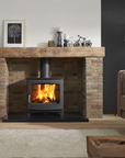 A black wood-burning fireplace with a glass front and active fire, set within a brick fireplace surround with a rustic wooden mantel in a living room setting.