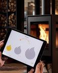 A person holds a tablet in front of a modern wood-burning stove, with a heating control app displayed on the screen.