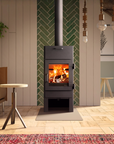A dark gray wood-burning stove with an open glass door showing a glowing fire, set against a green herringbone tile accent wall in a modern, cozy living room.