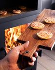 A hand holds a rectangular wooden pizza peel loaded with three small pizzas in front of an open wood-burning stove with visible flames.