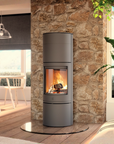 A modern cylindrical wood-burning stove with a visible fire, set against a rustic stone pillar in a contemporary living room interior.