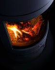 A close-up of a dark wood-burning stove with glowing orange and yellow flames visible through a glass front door, set against a black background.