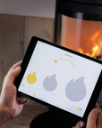 A person holds a tablet displaying a fireplace control app for a wood burning stove, with a real fireplace in the blurred background.