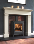 A cream-colored fireplace mantel and matching fireplace surround with a black wood-burning stove insert, set against a dark brick interior and a dark gray wall. Three lit candles sit on the mantel above the stove.