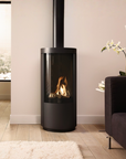 A modern black cylindrical wood-burning fireplace with a visible flame, set in a minimalist living room with light wood floors, a beige wall, and a dark gray sofa.
