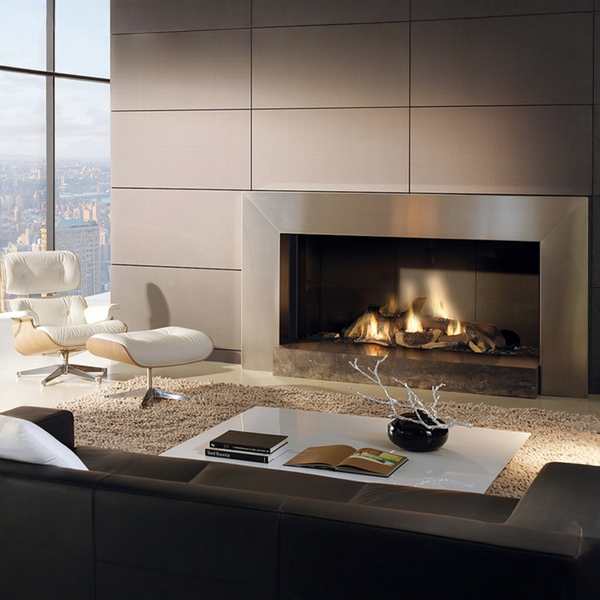 A modern living room interior featuring a large built-in gas fireplace with a metallic surround and visible flames, set against a paneled wall. A white lounge chair and ottoman sit to the left near a large window with a city view, and a dark sofa, coffee table, and decor are in the foreground.