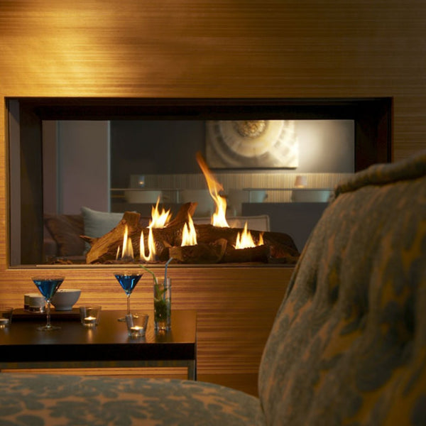 A modern gas fireplace with visible flames and logs, set in a warm wood-paneled interior with a blurred upholstered chair in the foreground and small table decor below.