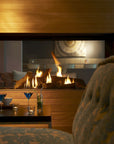 A modern gas fireplace with visible flames and logs, set in a warm wood-paneled interior with a blurred upholstered chair in the foreground and small table decor below.