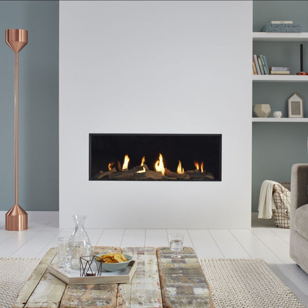 A modern linear gas fireplace with glowing orange and yellow flames set into a black interior, mounted on a white wall in a styled living room with a rustic wooden coffee table, a copper floor lamp, and built-in white shelves.