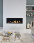 A modern wall-mounted linear gas fireplace with a black surround and visible flames, set into a white wall in a styled living room with a wooden coffee table, sofa, shelving, and a copper floor lamp.