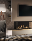 A modern gas fireplace installation in a styled living room, featuring a built-in linear fireplace with a low white hearth and active flames, a large wall-mounted TV, a gray sofa with a patterned throw pillow, and a lit floor lamp against patterned wallpaper.