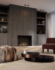 A modern living room interior featuring a large dark wood gas fireplace wall with built-in shelves, a gray sofa with a beige throw blanket, a dark red armchair, and two round black coffee tables on a beige rug.