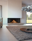 A modern living room featuring a built-in linear fireplace with a glowing fire, a large shag rug, a round wooden coffee table, a tufted sofa, and a glass bubble chandelier.