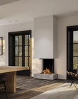 A modern minimalist interior featuring a sleek rectangular fireplace with a gray stone base, set against white walls with black-framed glass doors. A light wood dining table and a brown leather chair with a fur throw are visible in the foreground and a gas fireplace. 