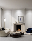 A modern living room featuring a cream curved armchair, a dark blue velvet accent chair, a round glossy coffee table, and a gas fireplace with a light stone mantel. The room has white walls with decorative molding, a large window with sheer curtains, and a light gray rug.
