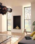 A modern living room interior featuring a black geometric pendant light hanging above a glass coffee table, a gray sofa with yellow pillows, a gas fireplace with a glowing log, and large windows looking out onto trees.