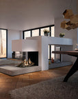 A modern open-plan living space featuring a contemporary white and stone gas fireplace with a glass front, set in a minimalist room with wood floors, a dining table, and a geometric gold pendant light.