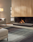 Modern gas fireplace wall in a styled living room, featuring a black-burning fireplace with a gray stone hearth, light wood cabinet doors above, a gray rug, and part of a beige sofa in the foreground.