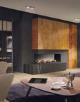 A modern living room interior featuring a black linear gas fireplace with a large wood-paneled surround, set against dark charcoal walls. In the foreground, a curved wooden coffee table holds a moka pot and cups on a dark purple rug, with a light beige armchair partially visible on the left.