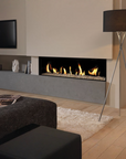 A modern linear gas fireplace with a black firebox and visible flames, set into a beige wall above a gray mantel in a styled living room. A black floor lamp, dark ottoman, and light wood flooring are also visible.
