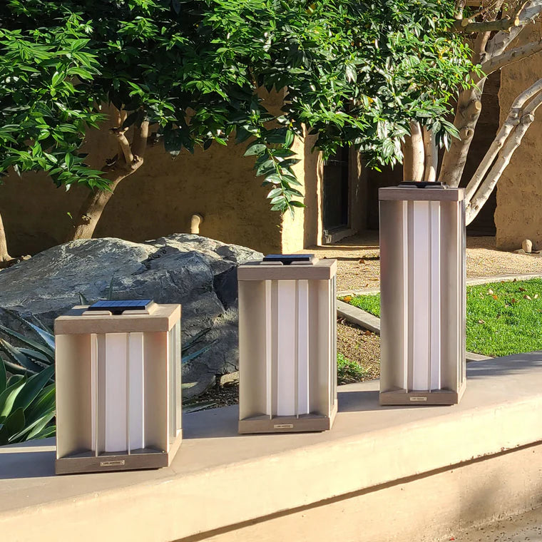 Three outdoor weathered teak solar lamps on a patio with greenery and a building in the background by Les Jardins available at Spacio retail stores in Mumbai & Kolkata. 