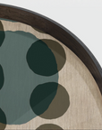 Close-up of a round decorative tray or plate with a dark wood rim, a beige textured base, and overlapping abstract dark green and taupe shapes on a light gray background.