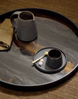 A round dark tray with a glossy marbled surface holding a gray ceramic pitcher, a matching cup and saucer with a small gold spoon, and a folded tan cloth, set on a brown textured surface.