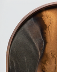 Close-up of a round decorative tray or plate with a warm metallic bronze-brown interior and a dark charcoal-black textured section, framed by a copper-toned rim against a light gray background.